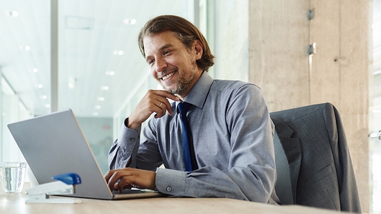 A man in a shirt and tie smiles while working on a laptop at a desk in a bright office.