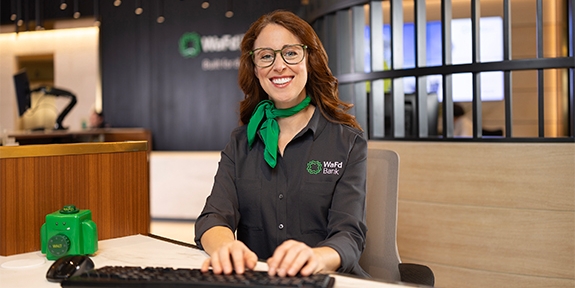 A smiling WaFd Bank representative in a branded shirt and green scarf sits at a desk, providing professional support to business clients.