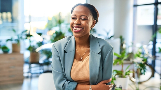 A professional Black woman in a blue blazer and peach top stands smiling in a bright, modern office surrounded by plants.
