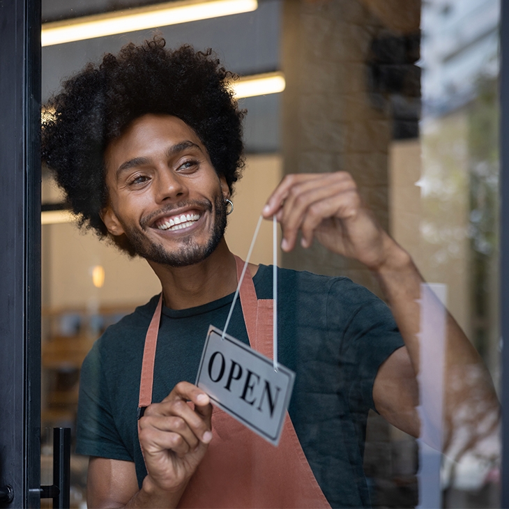 A smiling man with an afro hangs an "OPEN" sign on the glass door of his business.