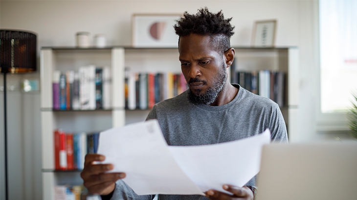 Serious looking man reading through paperwork with laptop open.