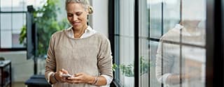 Person standing indoors and looking at a smartphone, with large windows and greenery in the background.