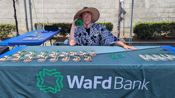 WaFd Bank employee sitting at table holding Walt the Vault piggy bank at Matsuri Festival.