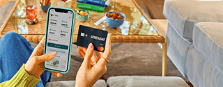 A person sitting indoors holds a smartphone in one hand displaying the Greenlight app with spending, saving, and investing balances, and a black Greenlight debit card in the other. A coffee table with snacks, books, and a drink is visible in the background, creating a casual home setting.
