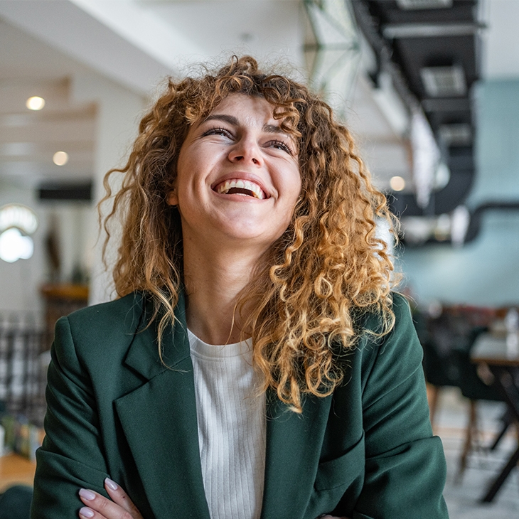 A happy, curly-haired woman in a green blazer is smiling and looking up while sitting in a cafe.