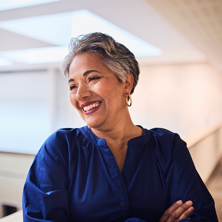 A smiling woman with short gray hair and a dark blue shirt stands with her arms crossed in a bright, modern indoor office space.