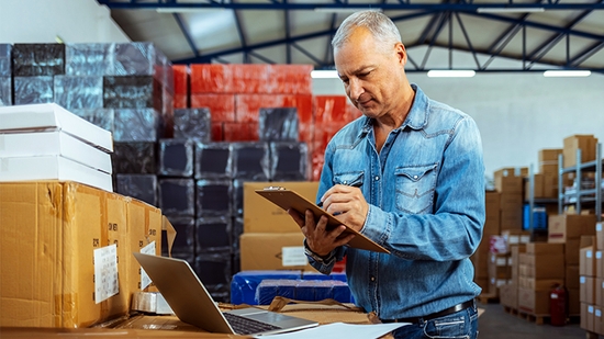 A man in a denim shirt stands in a warehouse full of stacked boxes, writing on a clipboard with a laptop open nearby.
