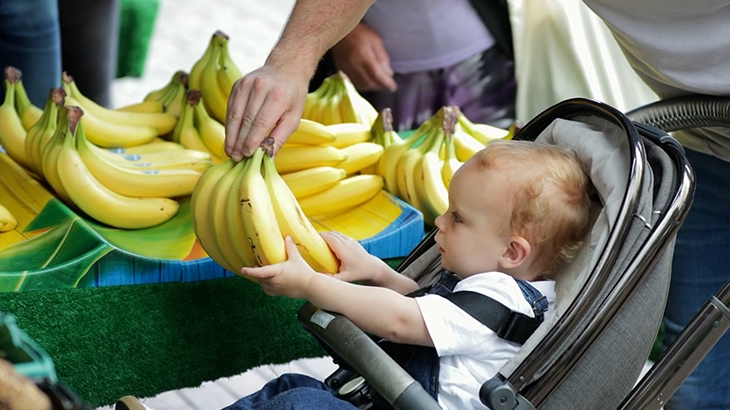 Baby in stroller reaching for bananas at a market.