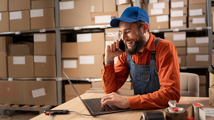 A smiling warehouse worker in a blue cap and orange shirt talks on a cell phone while using a laptop at a desk surrounded by cardboard boxes.