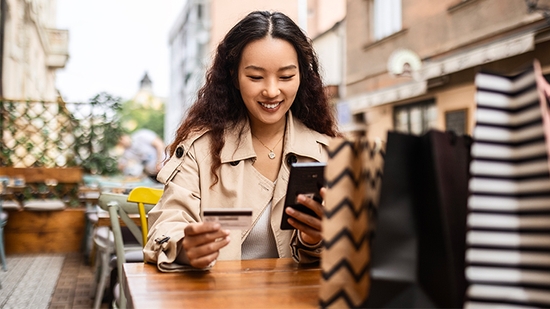 Woman holding credit card and mobile phone while at an outdoor cafe with shopping bags.