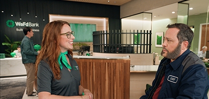 A smiling WaFd Bank employee wearing a green scarf talks to a male customer in a work jacket inside a WaFd branch.