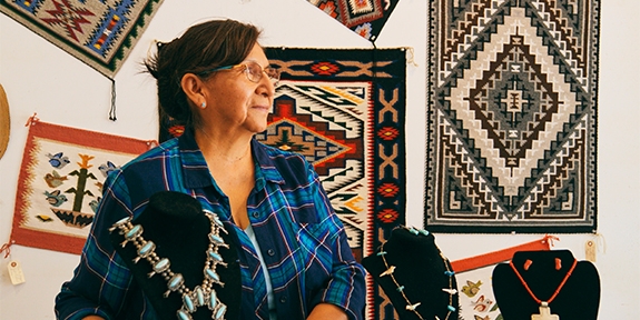 An older woman in a plaid shirt stands in a shop featuring Native American jewelry and woven rugs on the wall.