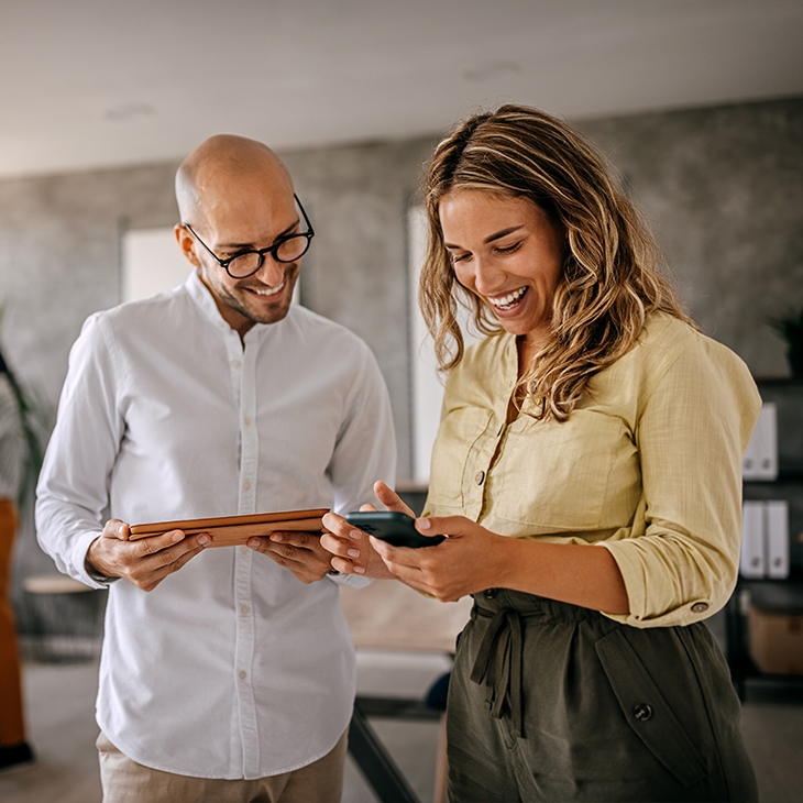 Two smiling professionals, a man and a woman, look at a tablet and a phone while standing in a busy, modern office.