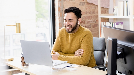 A bearded man in a mustard yellow sweater sits at a desk in a brick-walled office, smiling brightly at a laptop screen while wearing earbuds.