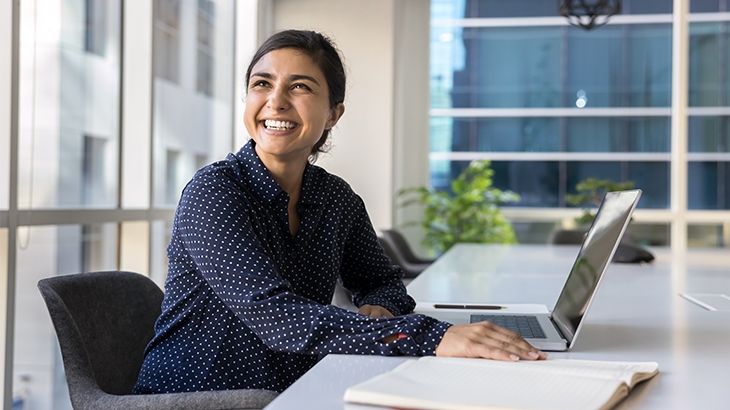 A professional woman in a navy blue, white-dotted blouse is smiling and looking up and away from her laptop while sitting at a conference table in a modern office.