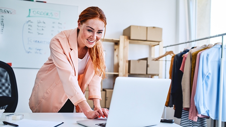 Smiling woman with reddish hair working on a laptop in a small business office surrounded by clothes on a rack and boxes.