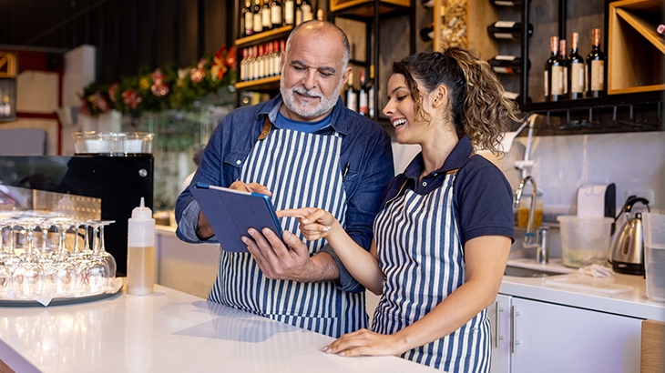 Two restaurant workers in striped aprons, one man and one woman, smiling while reviewing information on a tablet behind a counter.