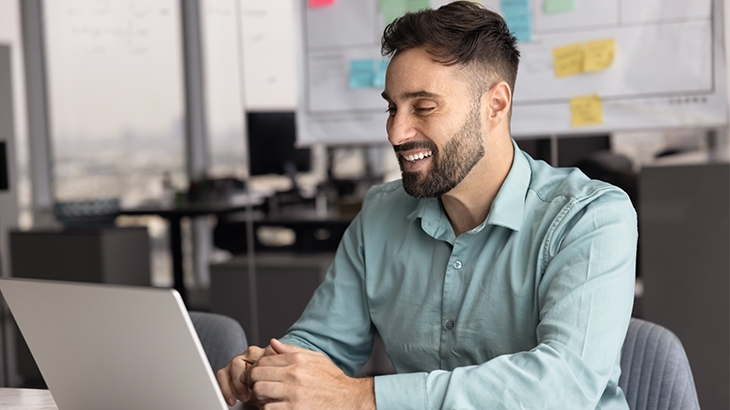 A bearded, smiling man in a light blue shirt works at a laptop in a modern office.