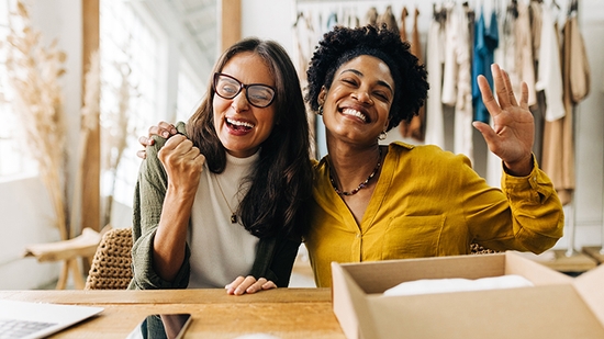 A diverse pair of happy businesswomen, smiling and gesturing enthusiastically in their clothing shop.