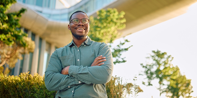 A smiling man with glasses and arms crossed stands outdoors in front of a modern glass building and a skybridge.