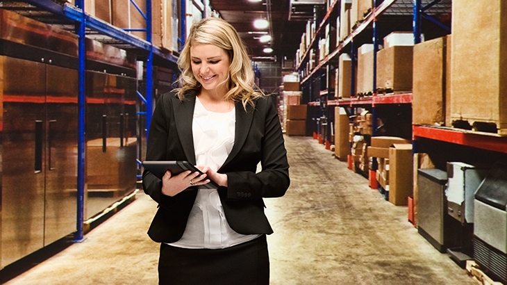A smiling Blonde woman in a business suit is standing in a warehouse aisle with tall metal shelving filled with cardboard boxes, looking down at and operating a digital tablet.