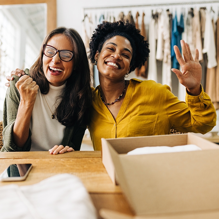 A diverse pair of happy businesswomen, smiling and gesturing enthusiastically in their clothing shop.