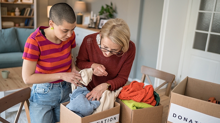 Two women, one younger with a shaved head and one older with glasses, look through boxes labeled "Donation" containing clothes in a home setting.