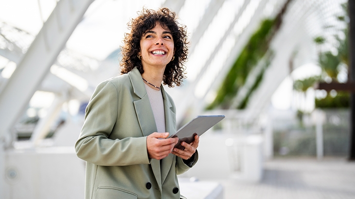A cheerful woman with dark, curly hair, wearing a light green blazer, stands outside holding a tablet and looking up and away with a wide smile.