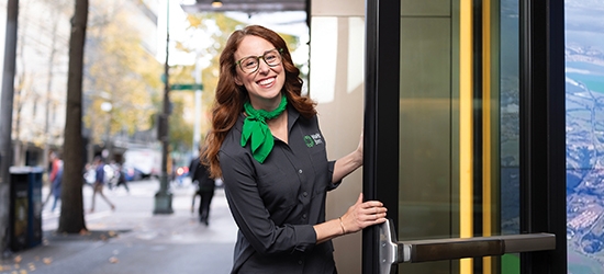 A smiling woman in a gray WaFd Bank uniform and green scarf holds open a glass door to a bank branch.