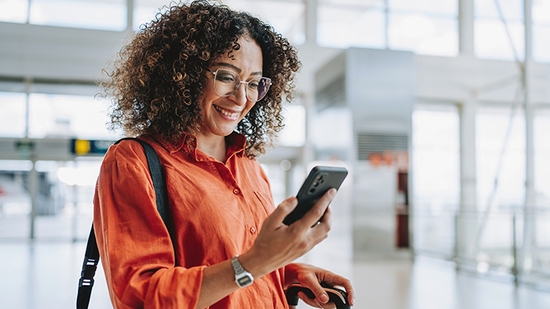 A smiling woman with curly hair and glasses looks at her smartphone while holding a suitcase in an airport terminal.