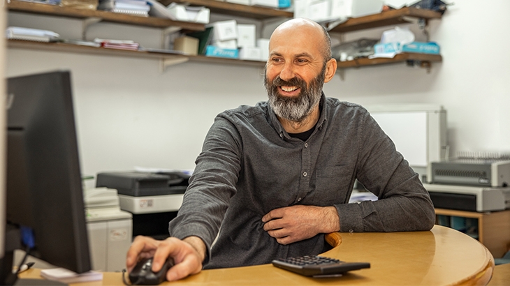 A bearded, bald man wearing a gray shirt is seated at a desk, smiling and looking at a computer monitor while operating a mouse.