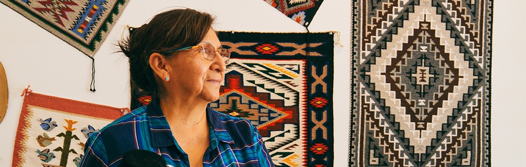 An older woman in a plaid shirt stands in a shop featuring Native American jewelry and woven rugs on the wall.