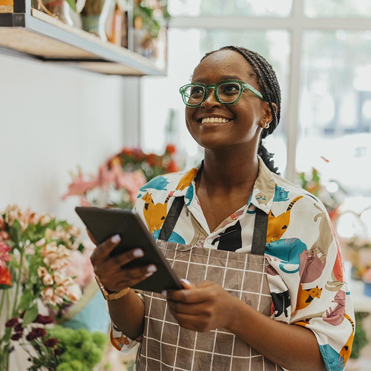 A cheerful Black woman with braided hair, wearing glasses and a patterned shirt under an apron, stands in a flower shop, holding a tablet and smiling.