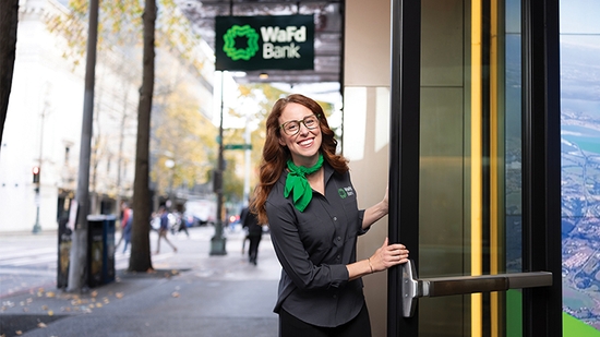 A smiling woman in a gray WaFd Bank uniform and green scarf holds open a glass door to a bank branch.