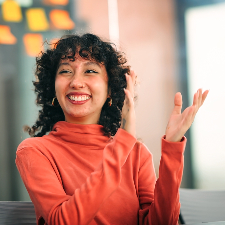 A happy woman with curly hair and a coral turtleneck is clapping in an office setting.