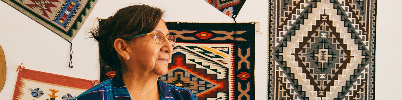 An older woman in a plaid shirt stands in a shop featuring Native American jewelry and woven rugs on the wall.