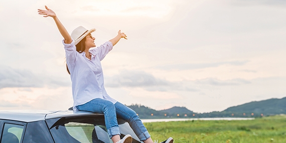 Woman sitting on a car with arms raised in celebration, looking at beautiful mountain scenery.