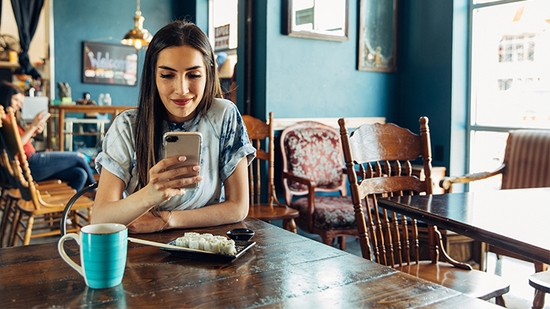 A woman sits at a wooden table in a cafe, looking at her smartphone while having a meal.