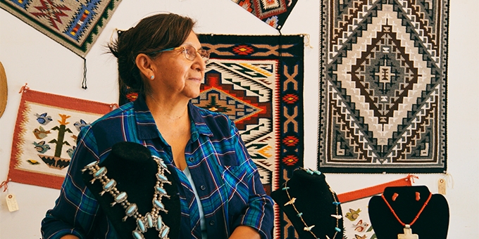An older woman in a plaid shirt stands in a shop featuring Native American jewelry and woven rugs on the wall.