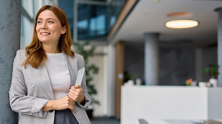 A confident, smiling woman in a light gray blazer, holding a tablet, stands in a modern, brightly lit office looking toward the window.
