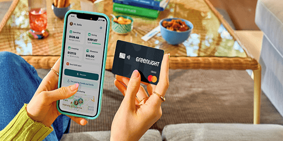 A person sitting indoors holds a smartphone in one hand displaying the Greenlight app with spending, saving, and investing balances, and a black Greenlight debit card in the other. A coffee table with snacks, books, and a drink is visible in the background, creating a casual home setting.