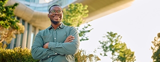 A smiling man with glasses and arms crossed stands outdoors in front of a modern glass building and a skybridge.