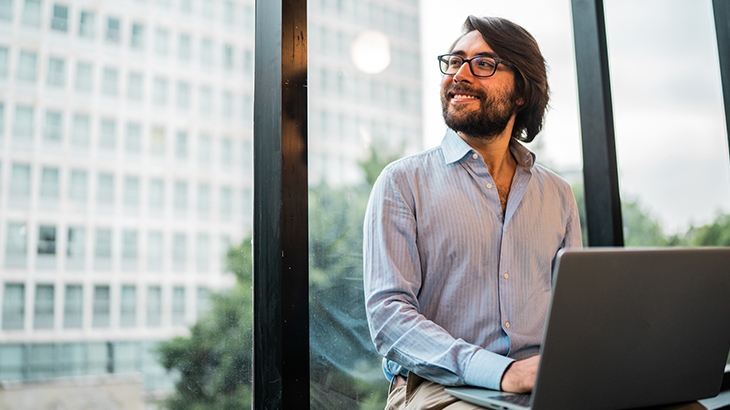 A man with glasses and a beard sits by a large window with a laptop, looking out at a city building.