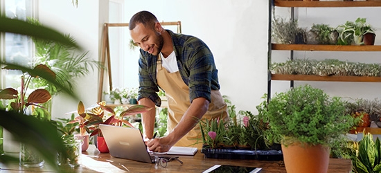 A business owner in an apron working on a laptop at a wooden table in a plant-filled shop or nursery.