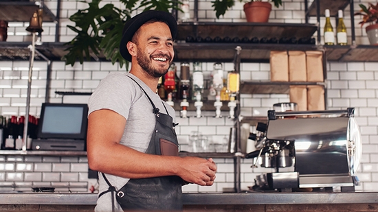 Smiling man in a black apron and fedora standing behind a counter with an espresso machine in a cafe.
