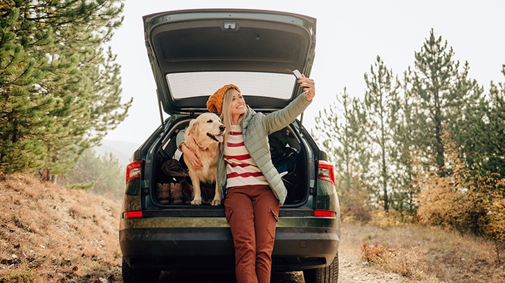 Woman taking selfie with dog sitting in trunk of car outdoors.