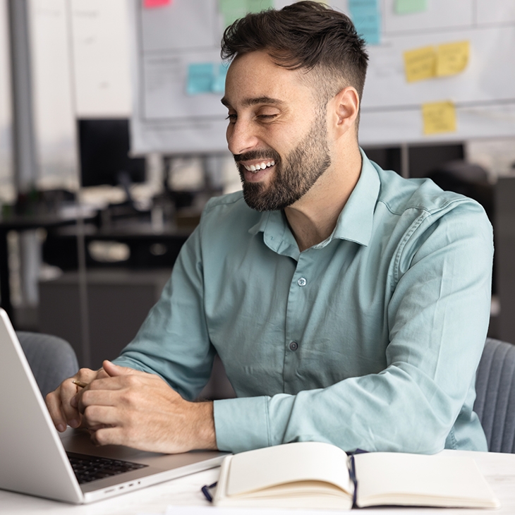 A bearded, smiling man in a light blue shirt works at a laptop in a modern office.