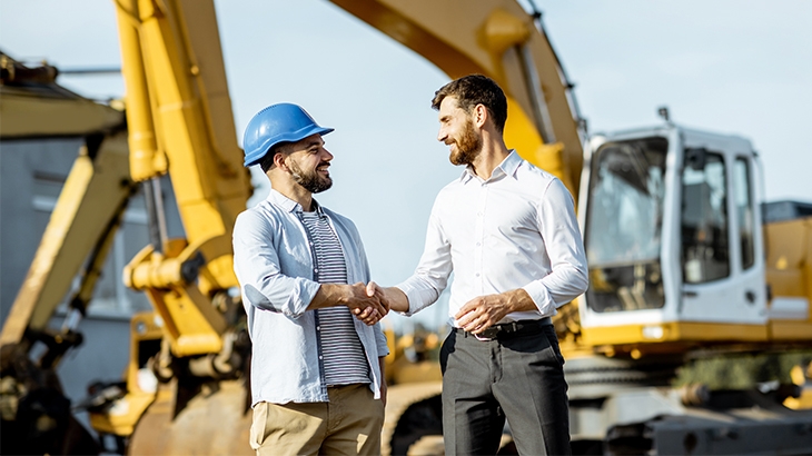 Two men shaking hands in front of heavy construction equipment.