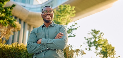 A smiling man with glasses and arms crossed stands outdoors in front of a modern glass building and a skybridge.