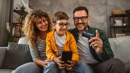 A happy family sitting together on a couch, with a child holding a smartphone and a man holding up a Greenlight card.